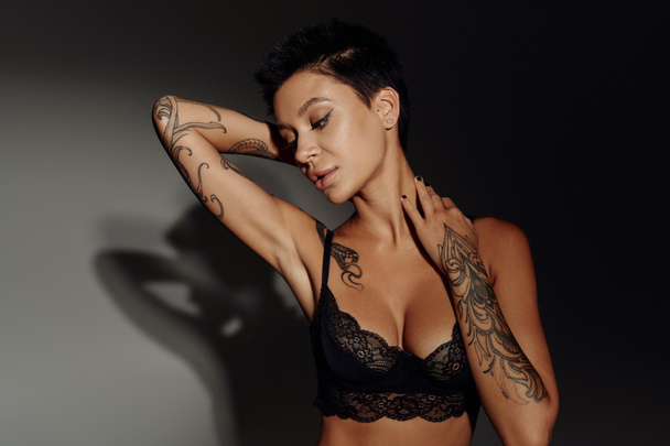 σέξι γυναίκα με τατουάζ σε δαντελένιο σουτιέν ποζάρουν σε μαύρο και γκρι φόντο με σκιά - Φωτογραφία, εικόνα