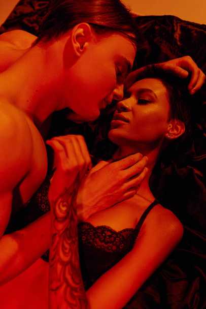 κορυφαία άποψη της γυναίκας με τατουάζ και νεαρός άνδρας που κάνει έρωτα σε μαύρο μεταξωτό κρεβάτι σε κόκκινο φως - Φωτογραφία, εικόνα