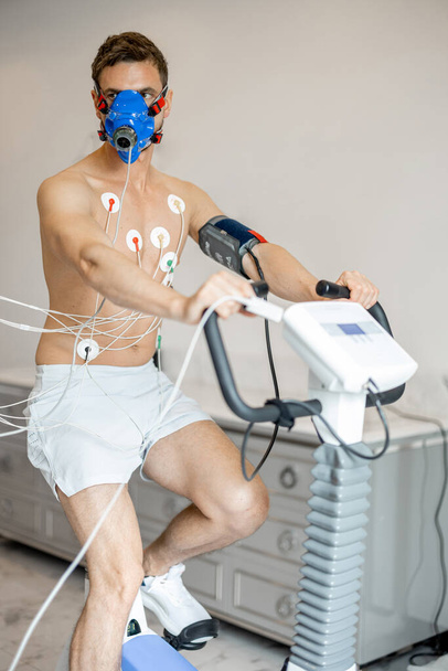Man athlete with breath mask and electrodes training on bike simulator, examining his cardiovascular system at medical office - Valokuva, kuva