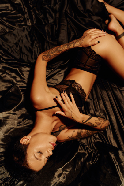 top view of tattooed woman with sexy body lying in lace underwear on black satin bedding - Zdjęcie, obraz