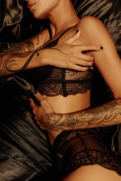верхний вид обрезанной татуированной женщины в сексуальном белье на черных шелковых постельных принадлежностях - Фото, изображение