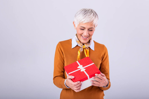 Happy mature woman in casual holding gift box and looking at it while enjoys over gray background. Senior woman hold gift in christmas style - Foto, Imagen