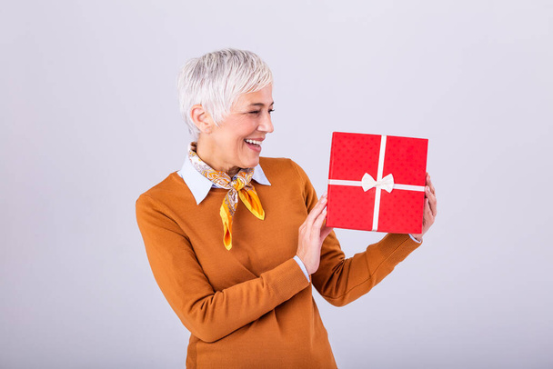 Portrait of cheerful positive glad charming aged woman with hairstyle having gift box in red package with white bow enjoying holiday festive mood isolated on grey background - Photo, Image