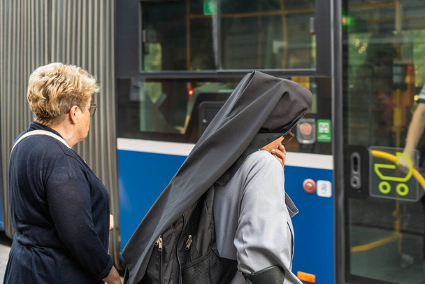 Catholic nun in monastic clothes stands thoughtfully at the door of a city bus at a public transport stop in Krakow, an ordinary woman behind a nun in line to enter the bus, two women - Foto, imagen