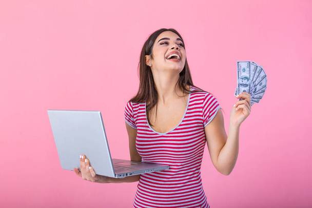 Portrait of excited satisfied girl holding money banknotes with laptop computer isolated over pink background. Portrait of a cheerful young woman holding money banknotes and laptop computer in hands - Foto, Imagem