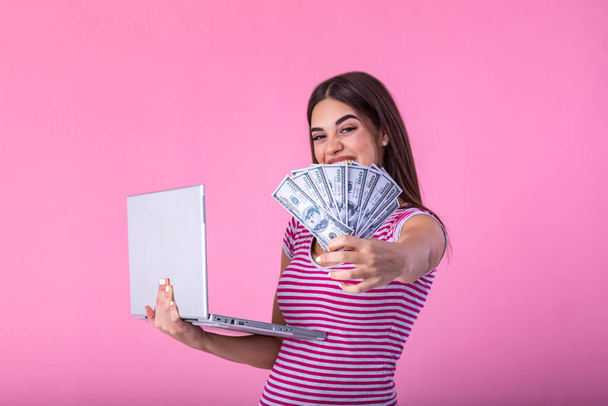 Portrait of an excited satisfied girl holding money banknotes with laptop computer isolated over pink background. Portrait of a cheerful young woman holding money banknotes and laptopcomputer in hands - 写真・画像