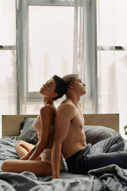 Homme torse nu en pyjama pantalon assis dos à dos avec petite amie sexy sur le lit  - Photo, image
