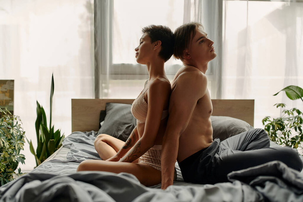 Sexy woman in lace lingerie sitting back to back with boyfriend in pajama pants on bed  - Fotografie, Obrázek