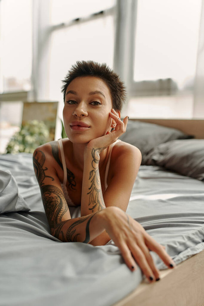 Sexy tatuado mulher olhando para a câmera enquanto deitado na cama de manhã  - Foto, Imagem