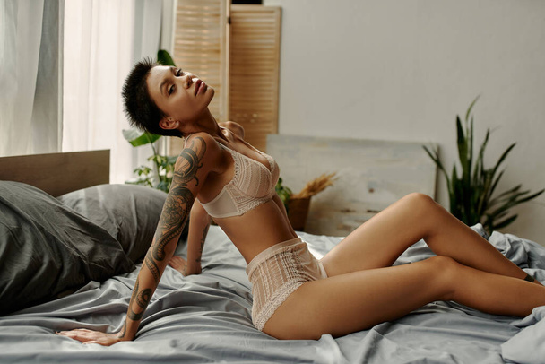 Tattooed woman in beige underwear looking at camera on bed  - Foto, imagen