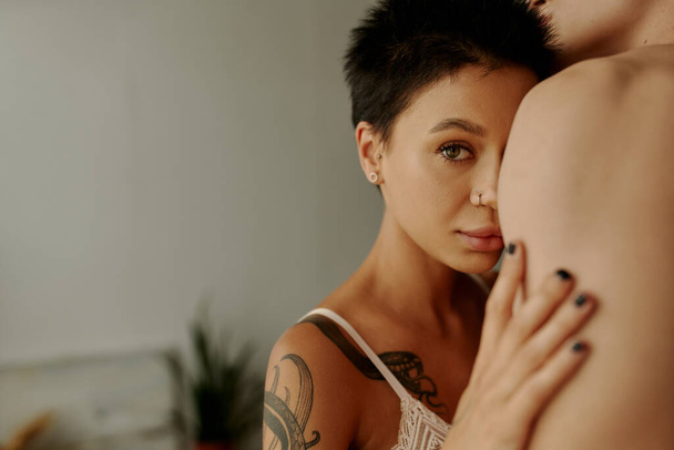 Tattooed woman in bra looking at camera near shirtless boyfriend at home  - Fotó, kép