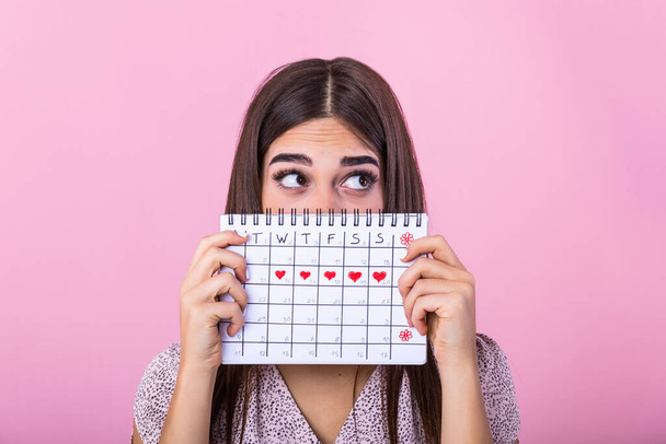 Portrait of a funny young girl in hiding behind a menstrual periods calendar and looking away at copy space isolated over pink background. Female Period calendar - Foto, Bild