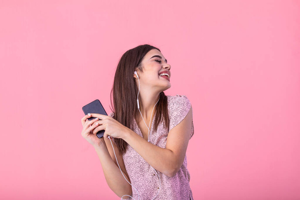 Усміхнена дівчина з коричневим волоссям насолоджується улюбленою піснею і танцями в рожевому верху. Крупним планом портрет збудженої молодої жінки, яка розважається в навушниках з телефоном на рожевому фоні студії
. - Фото, зображення