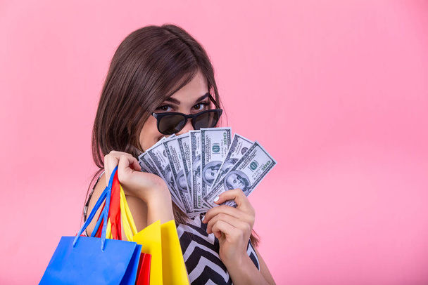 Shopping woman holding shopping bags in one hand and money in other looking at the camera on pink background. Beautiful young shopper smiling happy. - Photo, image