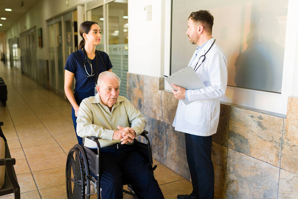 Hispanic nurse talking with the doctor at the retirement home while bringing a sick old man in a wheelchair at the nursing home - Фото, изображение