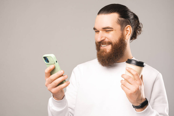 Close up portrait of a texting on phone man while holding to go cup of coffee. Studio photo over light grey background. - 写真・画像