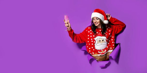 Happy Pretty brunette woman in red oversize pullover with Santa design peeking out of a hole in a purple studio paper background.  - Photo, image