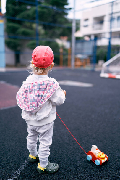Little girl is standing on the sports ground with a toy car. Back view. High quality photo - Foto, imagen