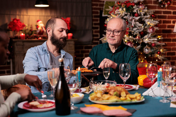 Diverse family having christmas festive dinner, eating traditional food at decorated table, man holding plate, passing dish. People gathering with parents on xmas holiday - Foto, Bild