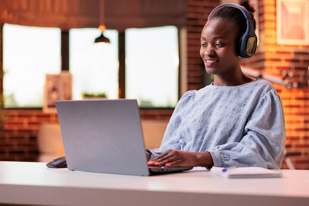 Smiling student in headphones attending online classes on laptop at home. African american woman watching educational video in modern room with big windows and beautiful warm sunset light - Foto, immagini
