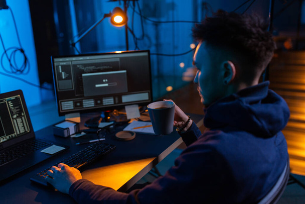 cybercrime, hacking and technology concept - male hacker in dark room drinking coffee and using computer virus program for cyber attack - Fotó, kép