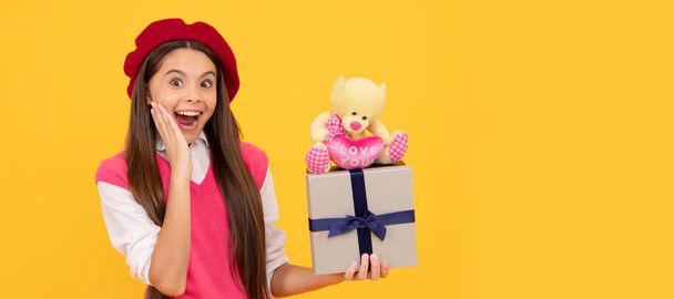 surprised teen girl in french beret hold toy present and gift box on yellow background. Kid girl with gift, horizontal poster. Banner header with copy space - Foto, Imagen