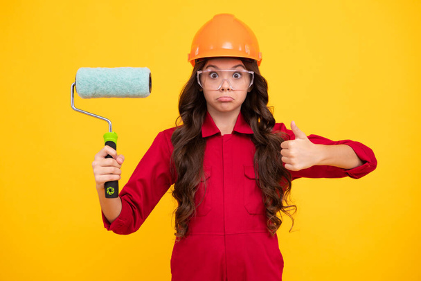 Funny face. Child builder in hard hat helmet. Teenage girl painter with painting brush tool or paint roller. Teenager worker isolated on yellow background. Kids renovation concept - Photo, Image