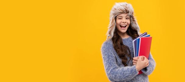 happy winking kid long hair in hat with copybook on yellow background, knowledge. Banner of child girl in winter hat, studio poster header with copy space - Photo, Image