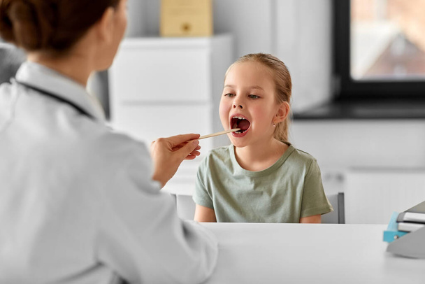 medicine, healthcare and pediatry concept - female doctor or pediatrician with tongue depressor checking little girl patients throat on medical exam at clinic - Foto, imagen
