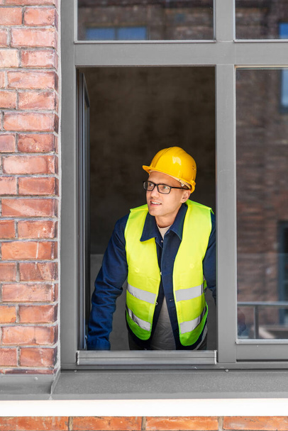 bouwbedrijf en bouwconcept - lachende mannelijke bouwer in helm en veiligheid west kijkend uit raam - Foto, afbeelding