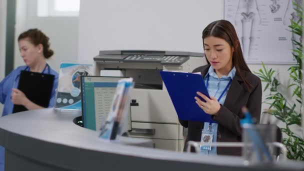Woman receptionist working on healthcare appointments with checkup report papers at hospital reception desk. Using consultation forms to give insurance support and help with examinations at counter. - Video, Çekim