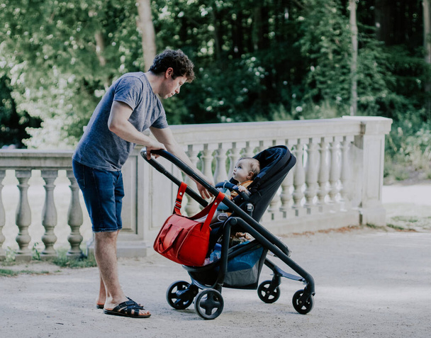 Caucasian young and handsome male dad pulls his hand to a little daughter sitting in a stroller and gently looking at him in a public park on the lawn near the lake, close-up side view. Concept of fatherhood, dads, family vacation, spring walk, dad c - Foto, Imagem