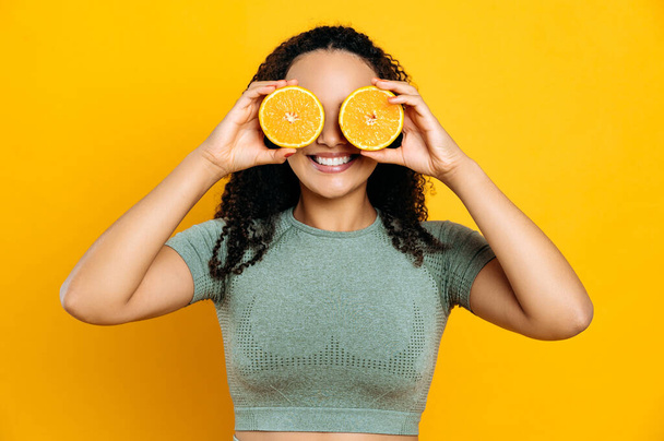 Healthy food and lifestyle, detox. Joyful lovely mixed race young curly haired woman, in sport outfits, standing on isolated orange background, holds two halves orange in her hands near eyes, smiling - Photo, image