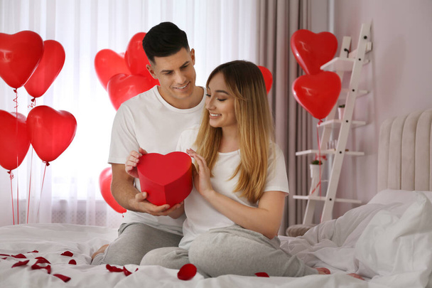 Man presenting gift to his girlfriend in room decorated with heart shaped balloons. Valentine's day celebration - 写真・画像