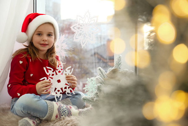 Little girl in Santa hat with paper snowflake near small Christmas tree at home - Foto, Bild
