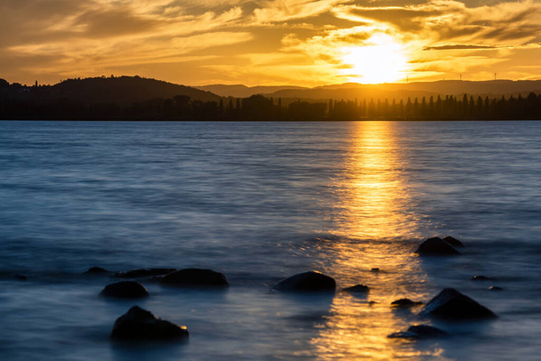 Золотой закат на Боденском озере с камнями на переднем плане и облаками в небе - Фото, изображение