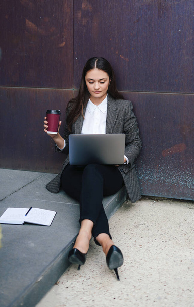Blanke vrouw met jas zittend op de vloer van de straat leunend op de muur van het kantoorgebouw met een videochat op laptop. Beste plek om op afstand te werken in - Foto, afbeelding