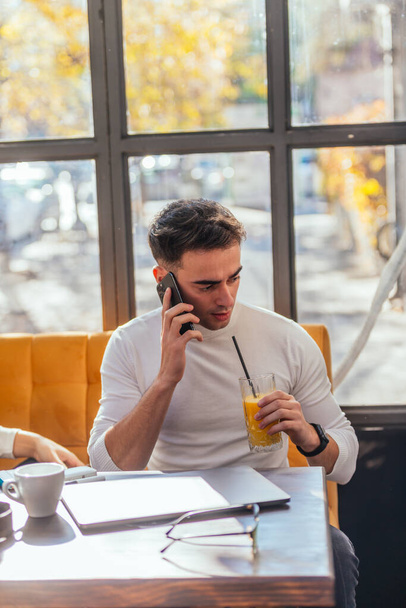カフェの窓の近くに座っている間、スマートでカジュアルな服装の若い男が携帯電話で話しています - 写真・画像