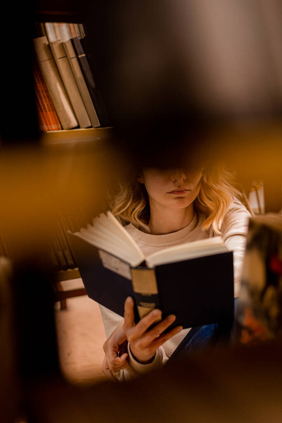 Девушка читает между книжными полками в библиотеке, сидя - Фото, изображение