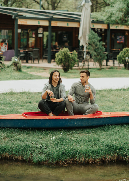 Pretty couple talking while sitting on a kayak in the park drinking orange juice - Fotó, kép