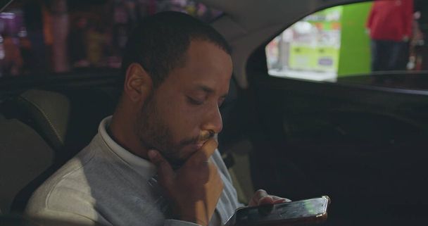 Person looking at cellphone riding taxi in backseat of car. African American person staring at smartphone screen after work in city - Photo, Image