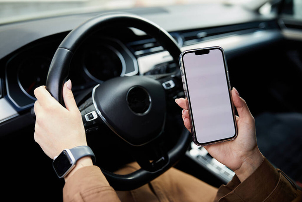 Primer plano de la Mujer sosteniendo el teléfono móvil negro en las manos con la pantalla de escritorio en blanco mientras el coche conduce en primavera en la ciudad. Imagen burlona - Foto, imagen