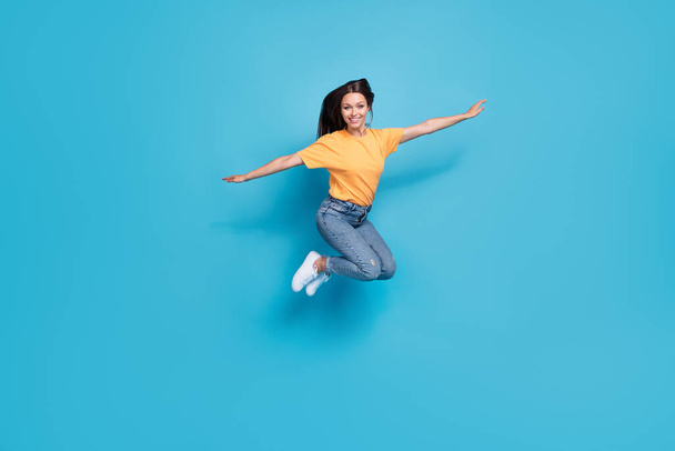 Full length photo of adorable pretty girl dressed yellow t-shirt arms sides jumping high isolated blue color background. - Foto, immagini
