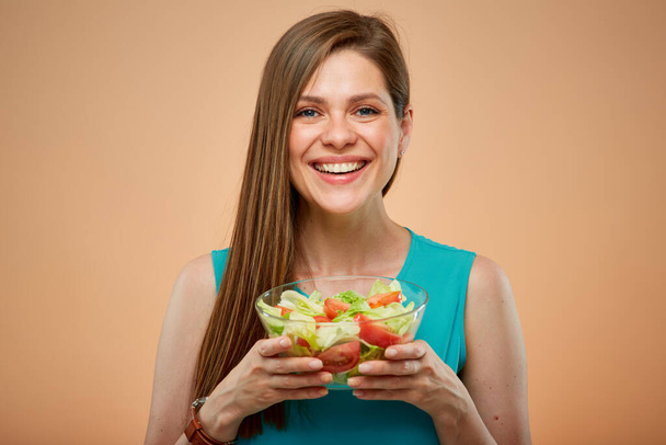 Young lady in azure dress holding green salad in glass bowl, isolated portrait on peach color background - Foto, Imagen