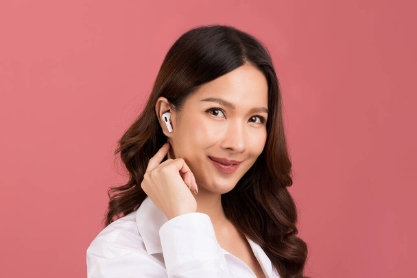 Long hair asian woman in wireless headphones white color listening to music isolated on pink background. - Zdjęcie, obraz