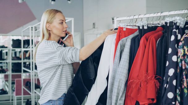 Female customer is slowly going through fashionable clothes on hanger in spacious shop. Other customers are moving around with lots of bright clothes and shoes in the background. - Foto, Bild