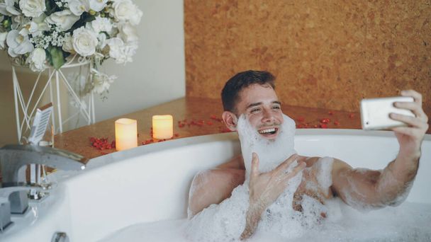 Cheerful young man with foam on beard is taking selfie using smartphone in hot tub in modern spa salon. He is laughing, gesturing, posing and looking at camera. - Foto, afbeelding