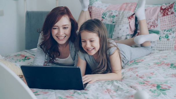 Happy cute daughter and young mother have online video call with father or grandmother using laptop computer lying on bed in cozy light bedroom at home. - Fotoğraf, Görsel