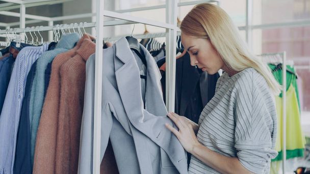 Close-up shot of young woman choosing coat in shop. First she is examining fabric carefully, looking at buttons, underlinimg and belt, then taking coat off hanger and taking it to fitting room - Фото, зображення