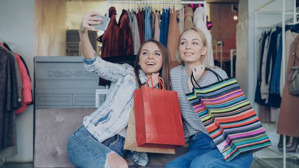 Cute girls are sitting and making selfie with colourful paper bags using smartphone in luxurious womens clothing boutique, then watching pictures together. They are smiling and laughing carelessly. - Фото, изображение